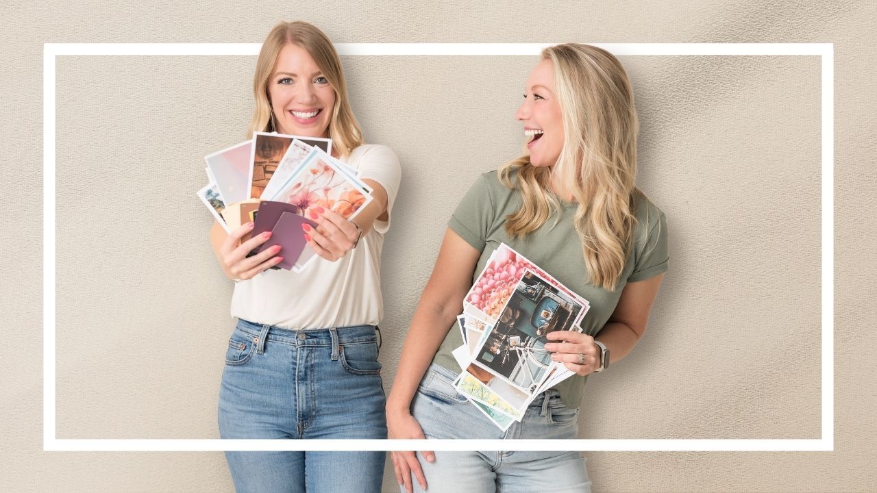 Two women laughing together while holding colorful magazines and printed photos against a neutral beige background