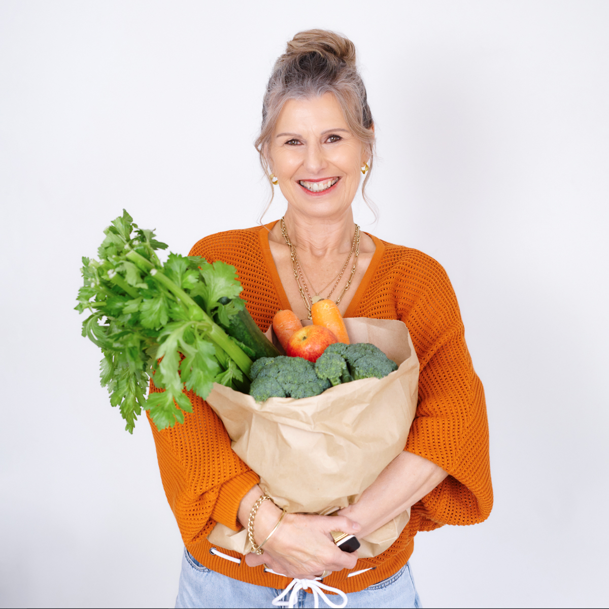 Ginny McArthur smiling in an orange jumper, holding a bag of fresh vegetables