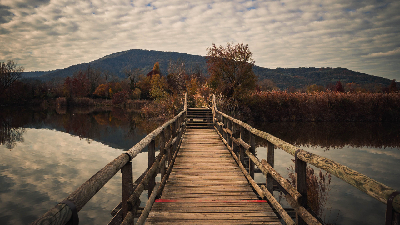 A wooden footbridge crossing a calm lake toward autumn trees and a distant hillside, evoking a season of pause and reflection before moving forward