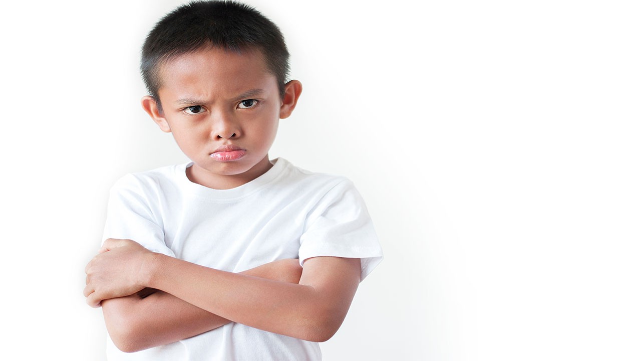 A young boy standing with his arms crossed and a defiant frown, looking angry.