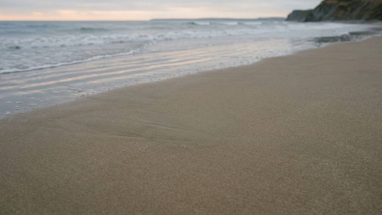 A peaceful, wide photograph of an empty sandy beach with soft waves receding under an overcast sky, leaving smooth, clear wet sand.