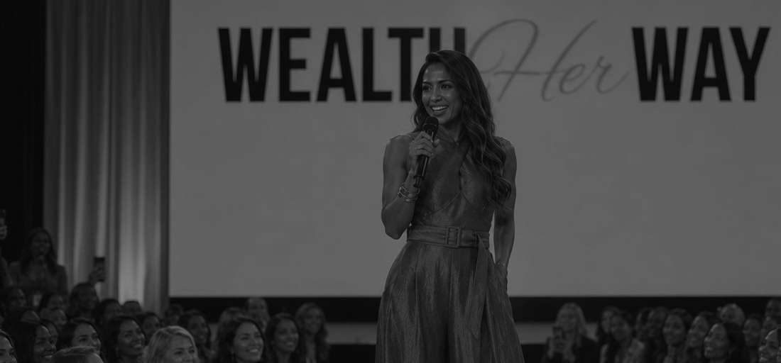 Black-and-white photograph of a woman speaking with a microphone on the Wealth Her Way stage in front of a seated audience.