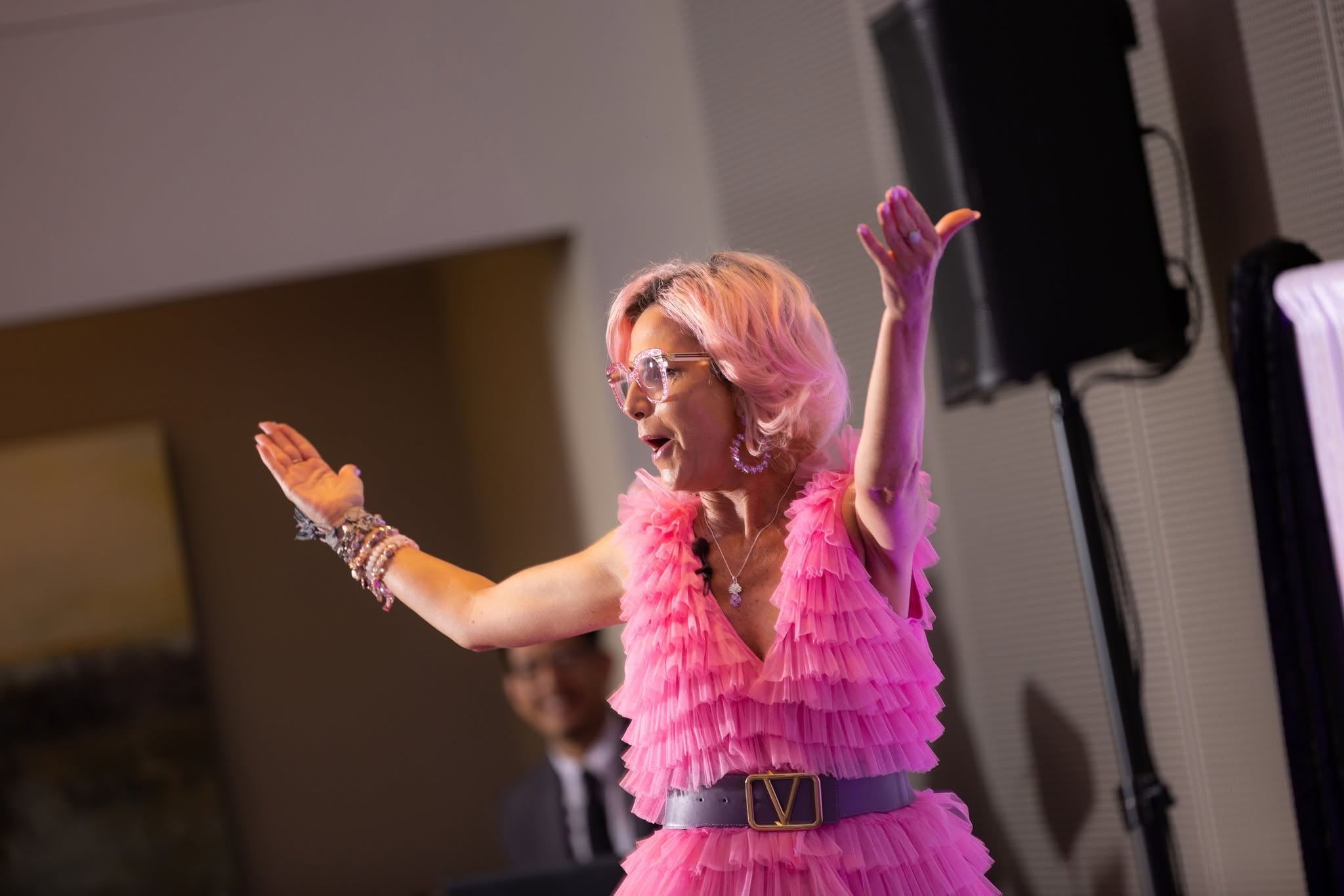 A speaker in a pink ruffled dress with her arms open, addressing the room.
