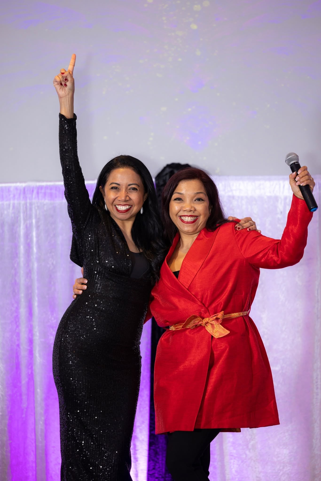 Two guests celebrating together in front of lilac-lit draping, one with her arm raised and the other holding a microphone.