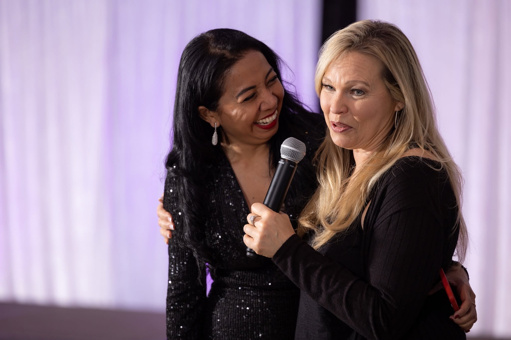 Two guests arm in arm in front of lilac-lit draping, one laughing as she holds a microphone.