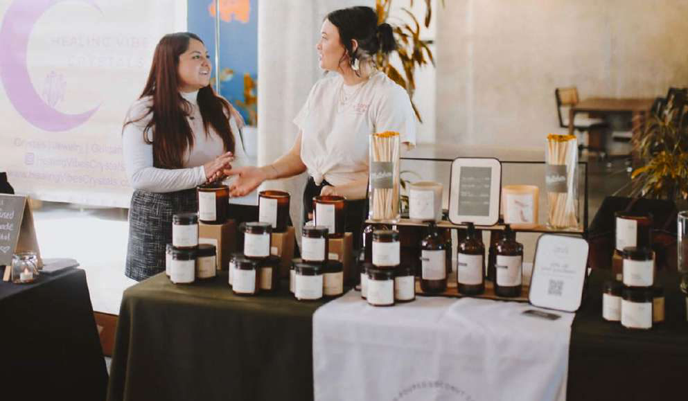 Two women talking behind a candle vendor table stacked with amber jars.
