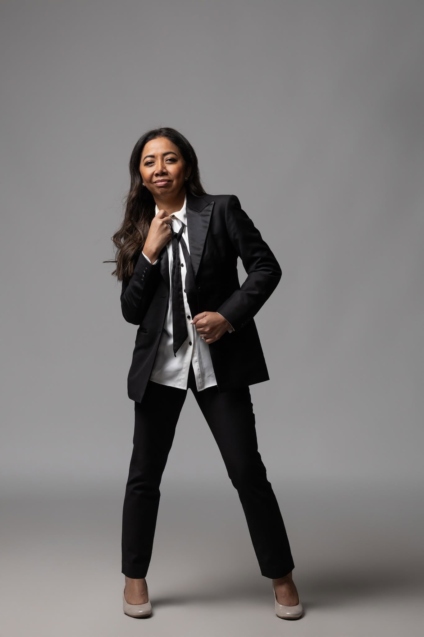 Nim Stant standing full length in a black suit and white shirt, photographed against a grey studio backdrop.