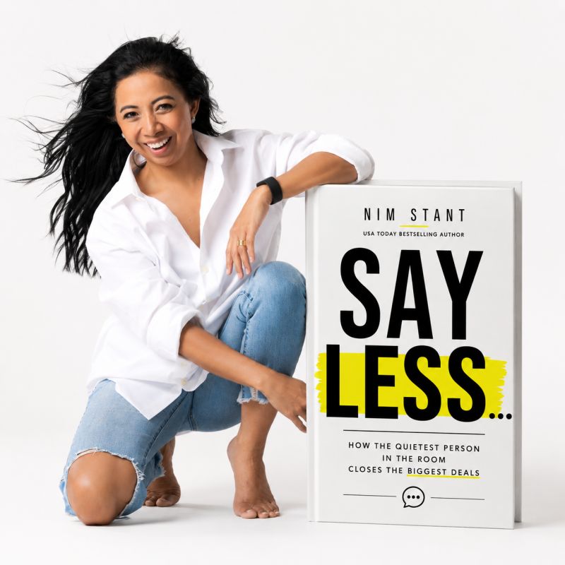 Nim Stant kneeling beside a large copy of her book Say Less… — How the Quietest Person in the Room Closes the Biggest Deals — with a yellow highlighter stroke through the word LESS.