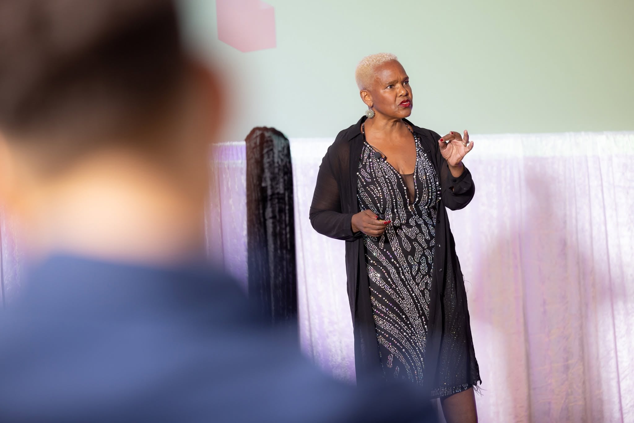 A speaker in a sequinned dress and sheer jacket addressing the room from the stage, framed past a guest's shoulder.