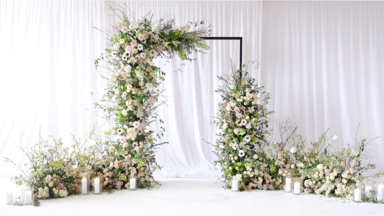 A floral archway with soft white, blush and green flowers, surrounded by low meadow-style arrangements and candles against white draping. From The Installations Masterclass