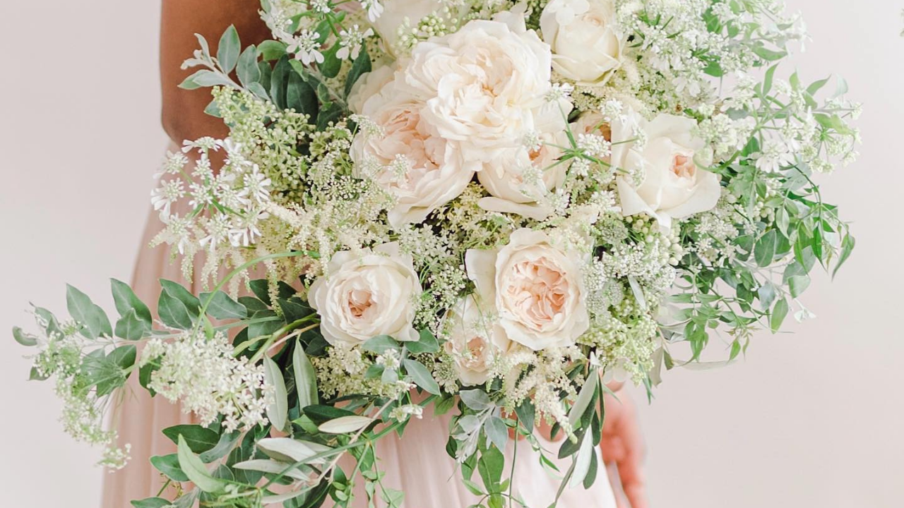 Soft, romantic bridal bouquet featuring ivory garden roses, delicate white umbels, trailing foliage and seasonal greenery in an airy garden style.