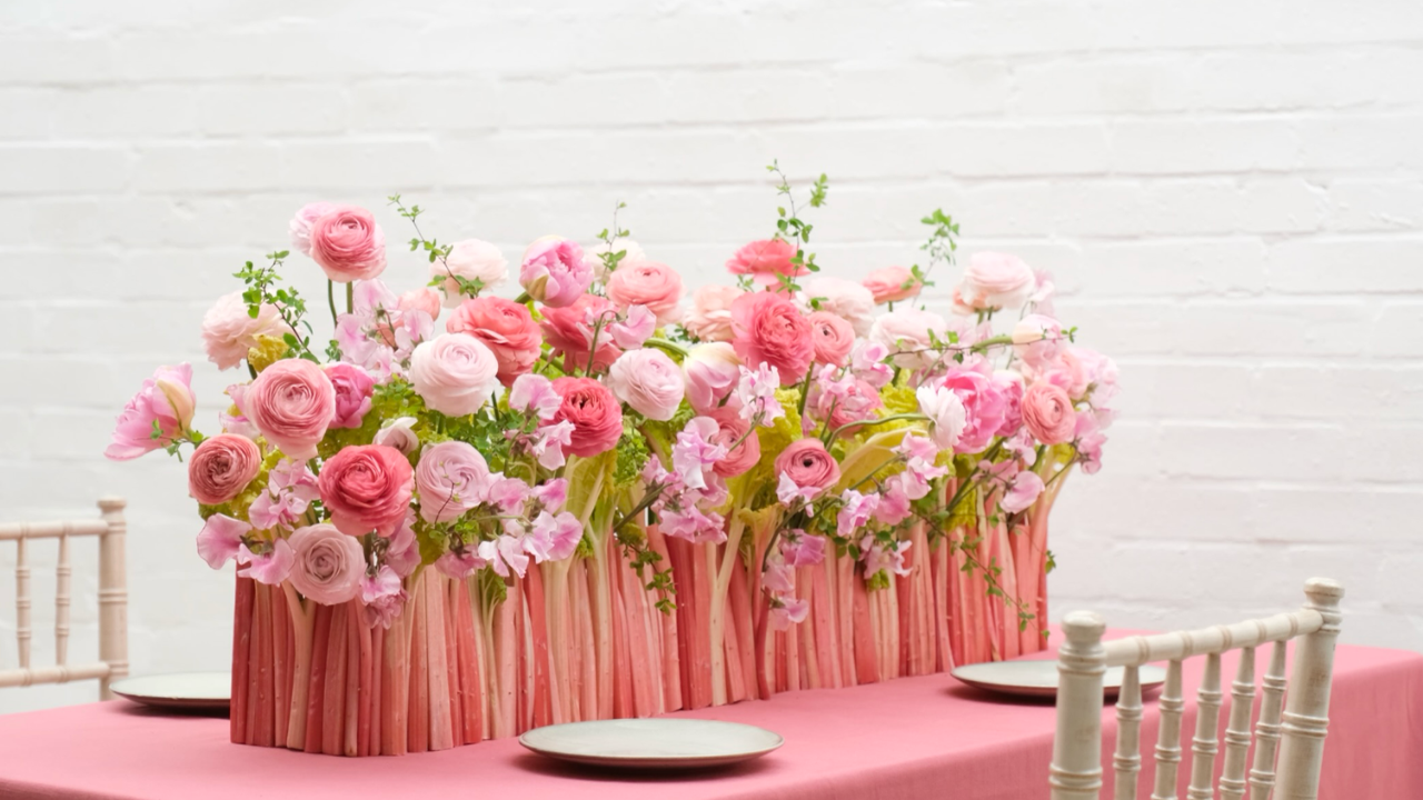 Pink floral centrepiece featuring ranunculus, sweet peas and sculptural rhubarb stems, arranged on a pink tablescape against a white brick wall.