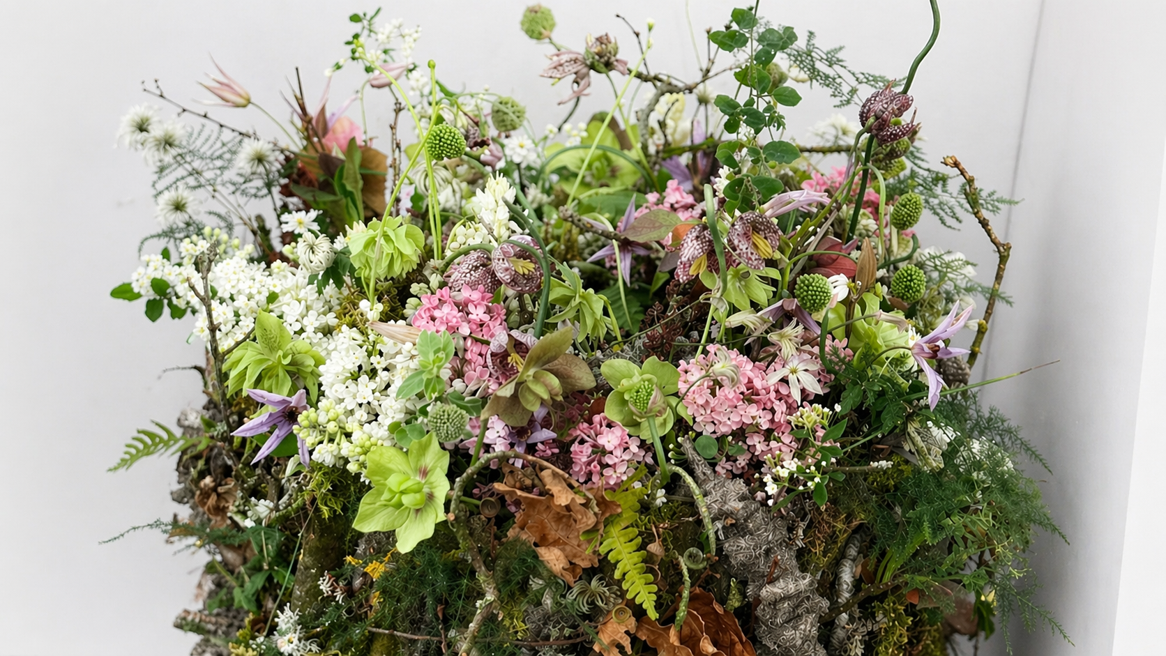 A natural, woodland-style floral arrangement composed of soft pink blossoms, pale green hellebores, delicate white flowers, and airy foliage, arranged amongst moss, branches, and textured greenery against a light, neutral background.