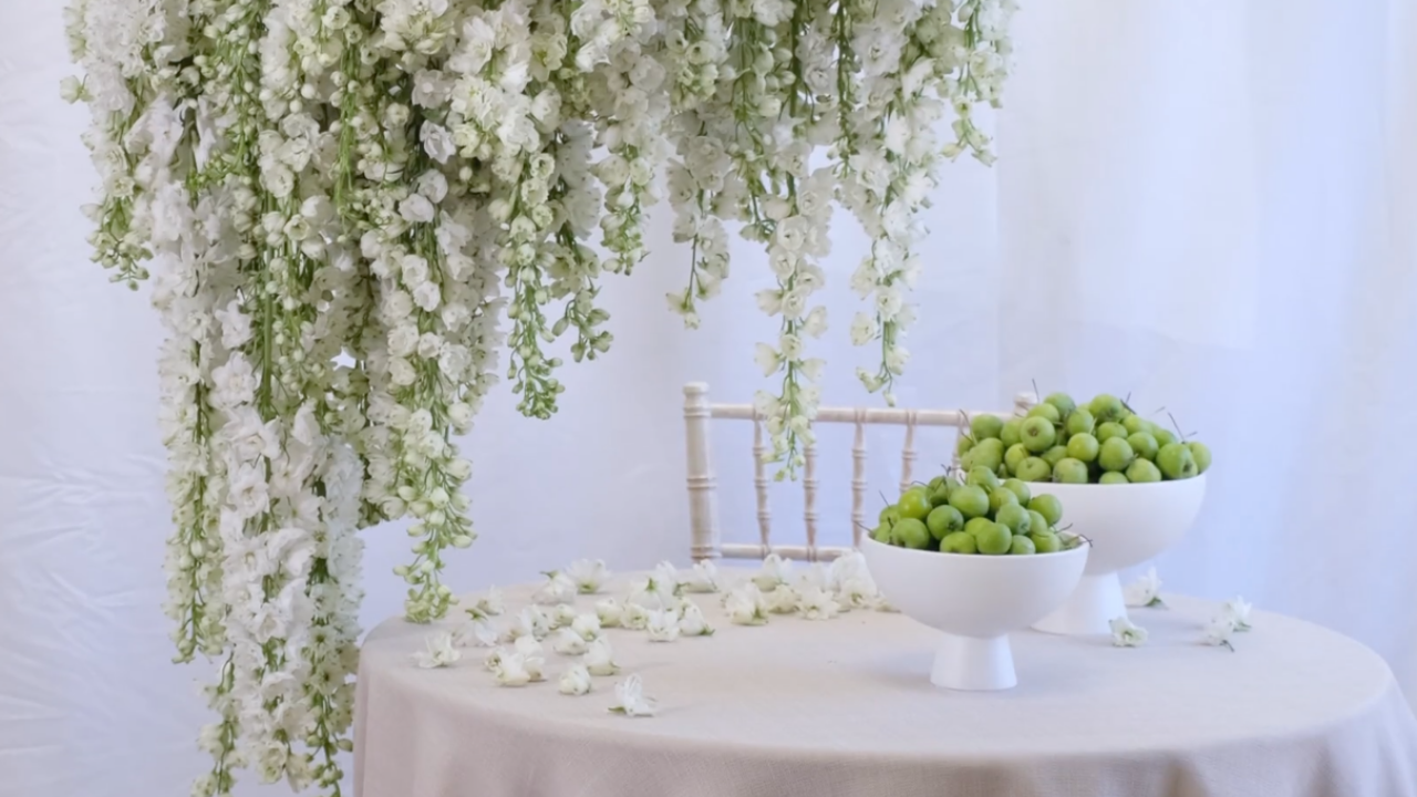 White floral wedding installation cascading above a softly styled table, with bowls of green apples and scattered white flowers creating a fresh, elegant tablescape.