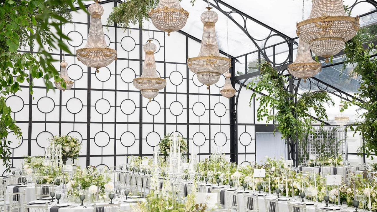 Luxury summer wedding reception inside a glass marquee, with white and green floral installations, hanging chandeliers and long flower-filled tables.