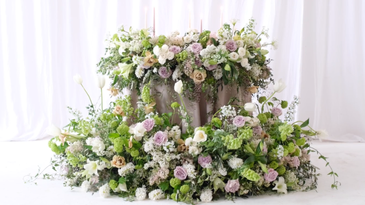 Lavish wedding flower installation with soft white, green, blush and lilac flowers arranged around a draped table, featuring roses, lilies, tulips and abundant seasonal foliage.