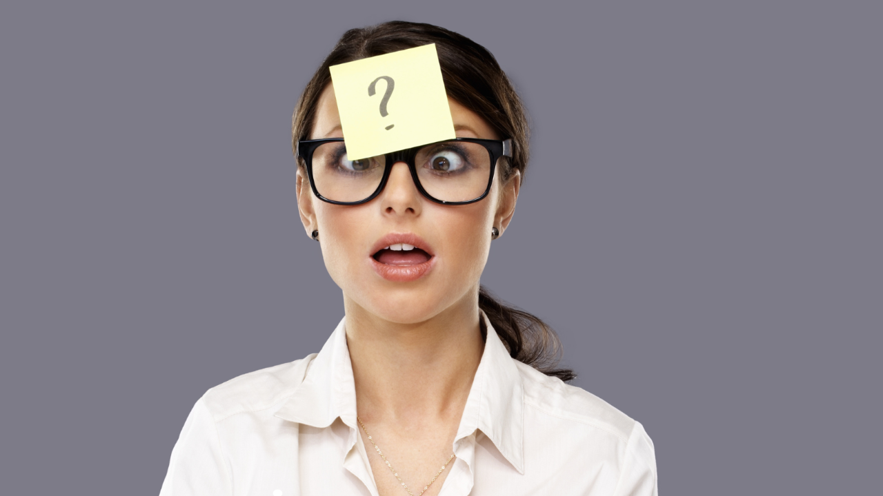 A woman with dark hair in a ponytail wearing a white shirt, black glasses against a grey background has a yellow post-it note with a question mark stuck to her forehead. 