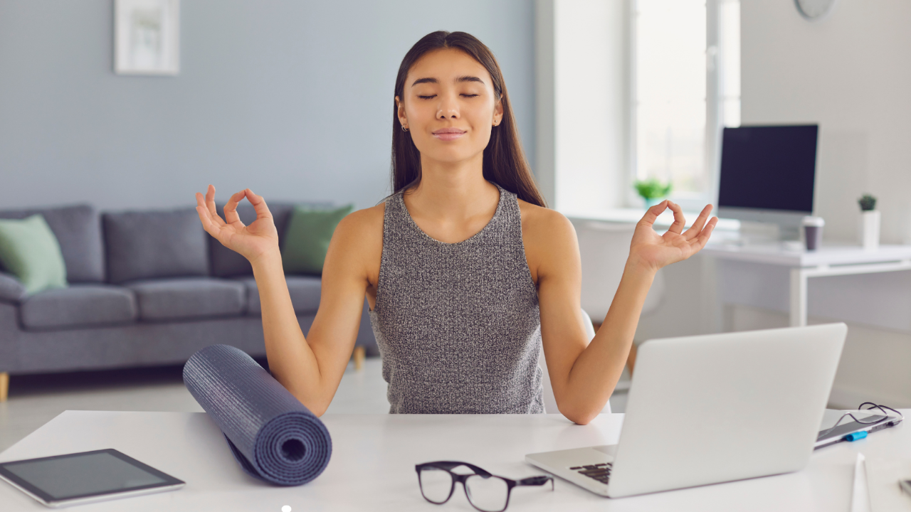 A woman with dark hair and a grey tank top sits at a desk with her hands out to her sides meditating. The white desktop has a blue yoga mat, tablet, glasses, and open laptop. 