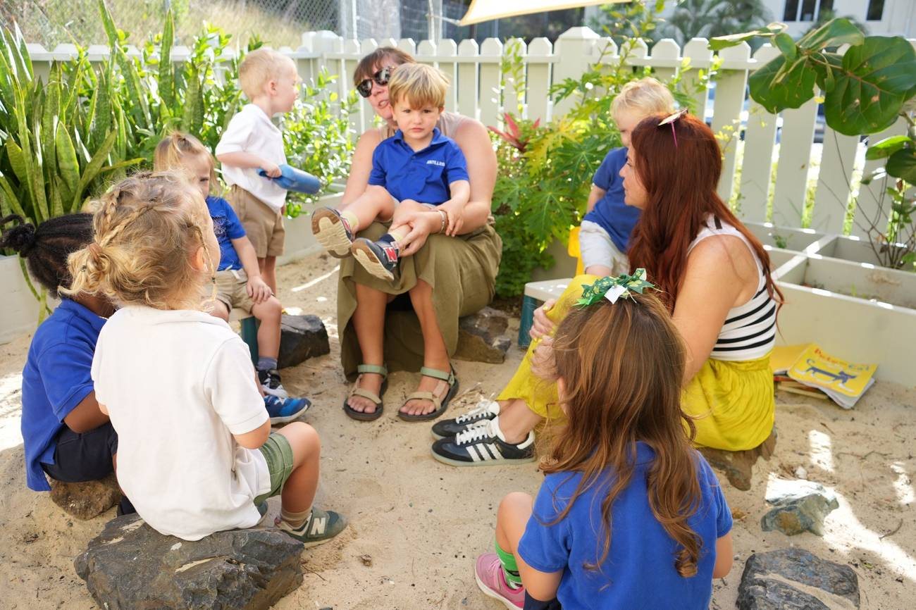 Kristen sitting in the sand outdoors with a circle of preschool children and another teacher