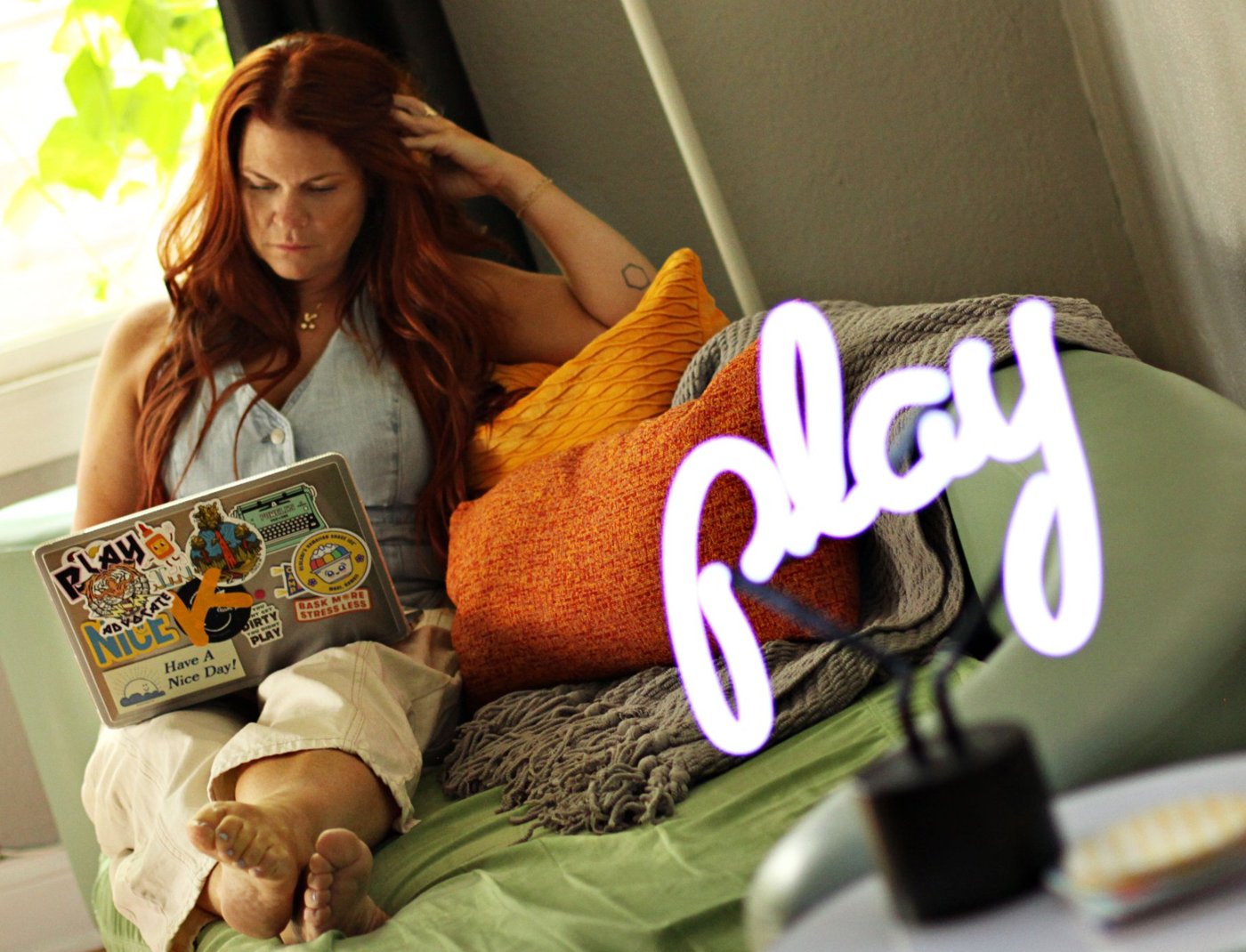 Kristen Day working on a sticker-covered laptop next to a glowing neon play sign