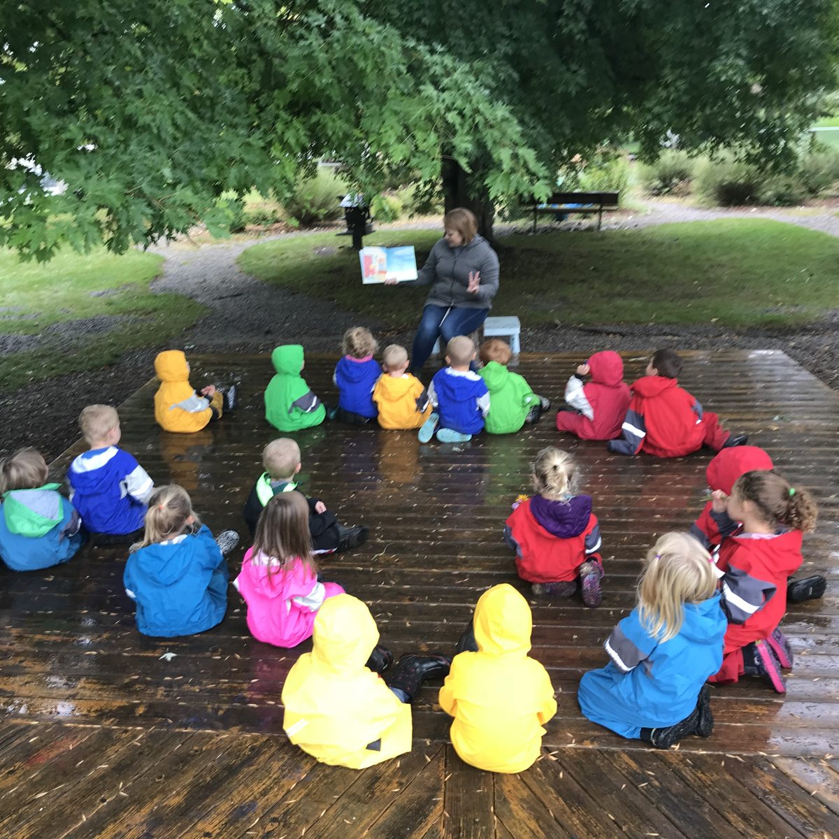 Kristen reading a picture book under a big tree to a circle of preschoolers in rainbow rain suits at Butterfly Hill Nature Preschool