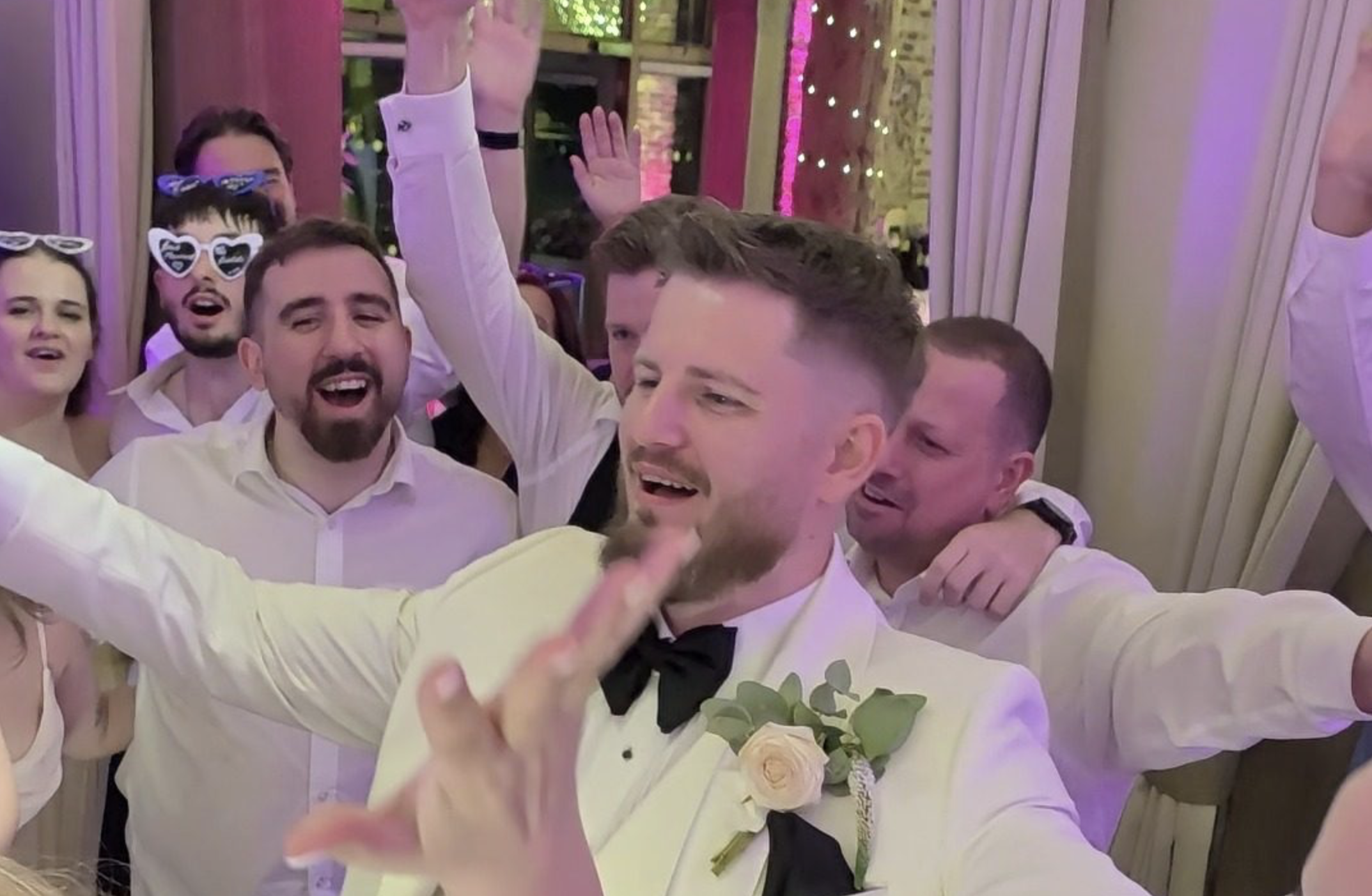 Groom singing with arms wide as guests cheer and raise a glass on a packed dancefloor