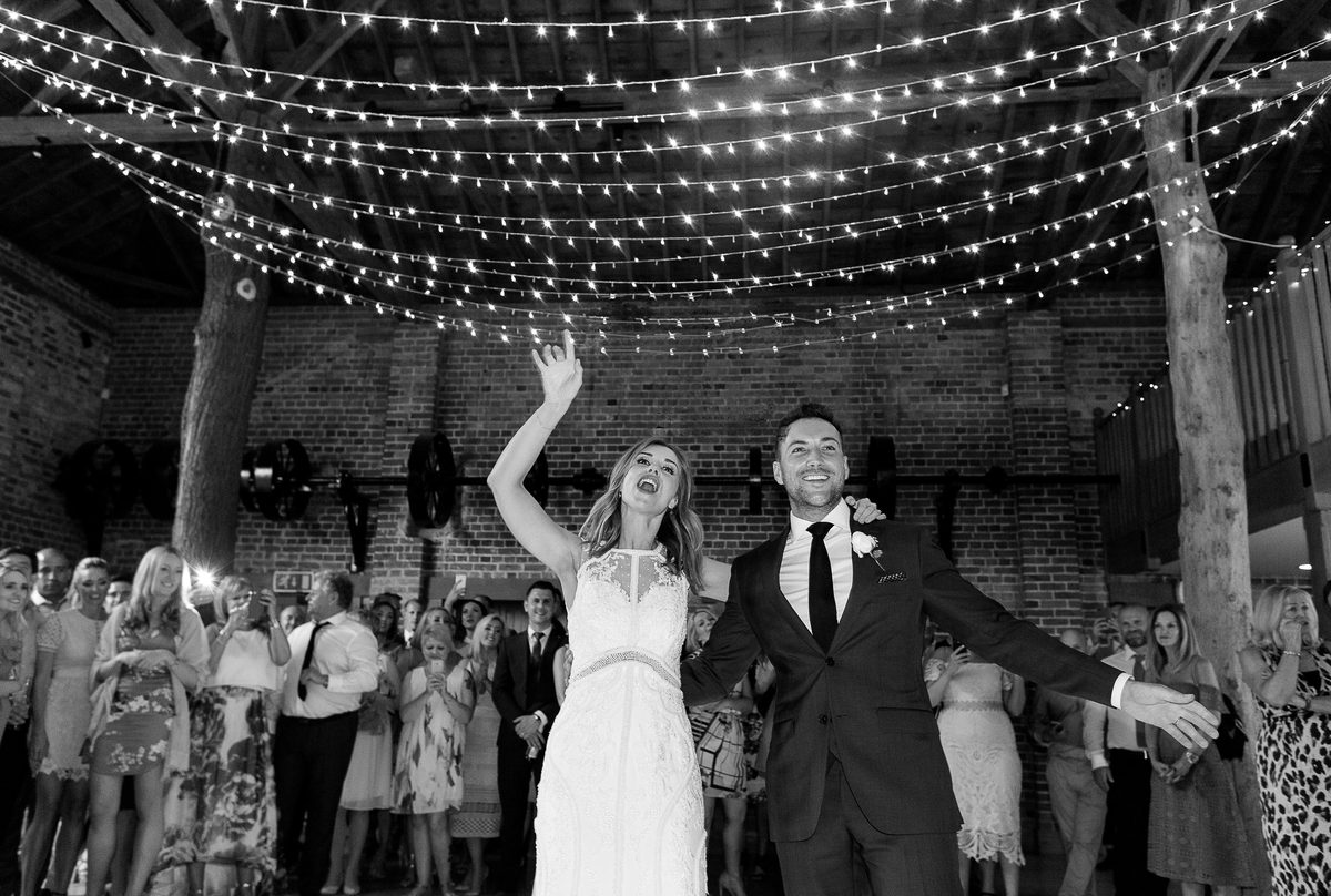 Bride singing with her arm raised beside the beaming groom under a canopy of fairy lights