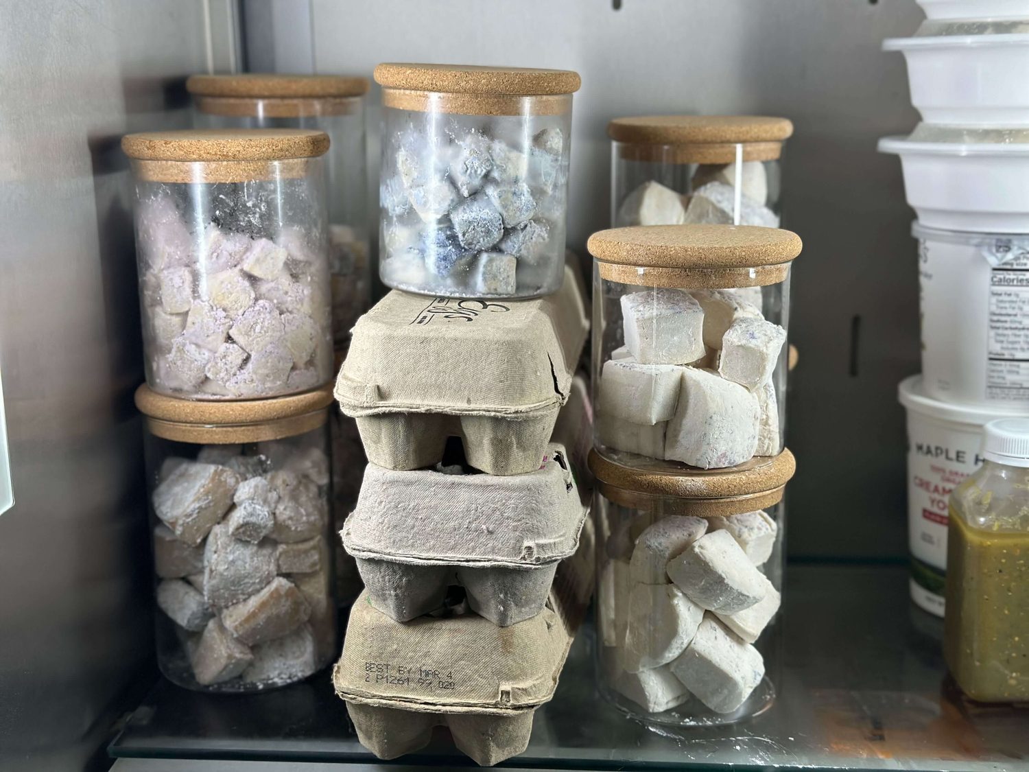 Glass storage jars with cork lids on a refrigerator shelf holding homemade marshmallows, beside egg cartons and plastic tubs