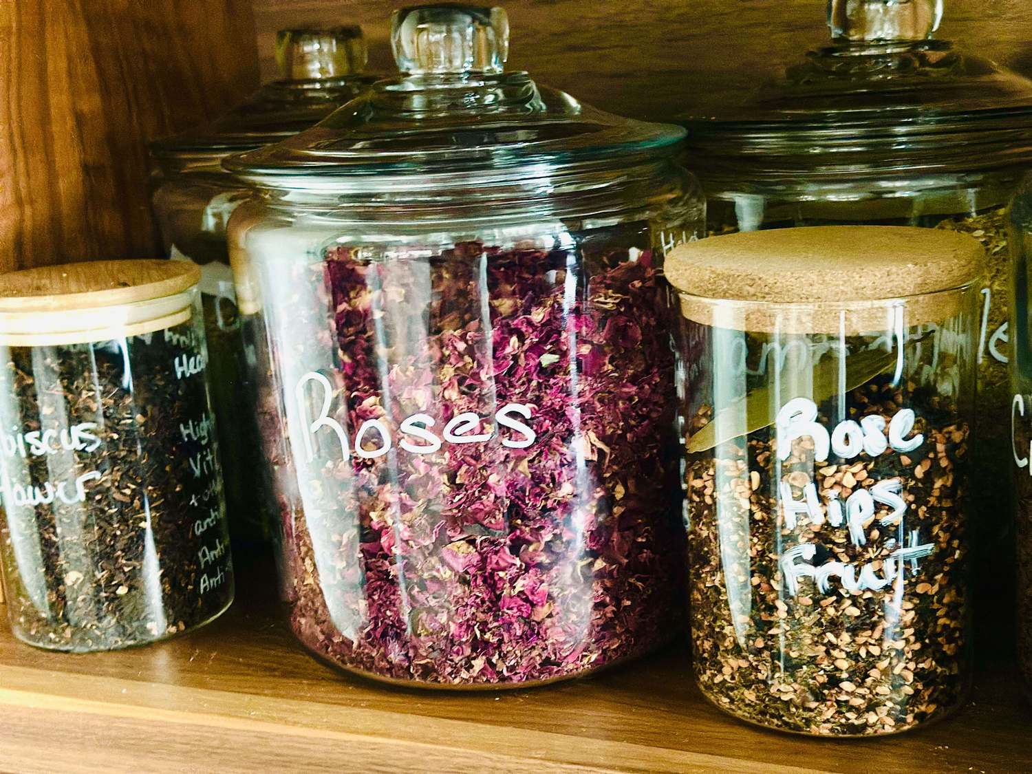 Glass apothecary jars with cork and glass lids holding dried roses, rose hips, and hibiscus flower, labeled in white chalk marker