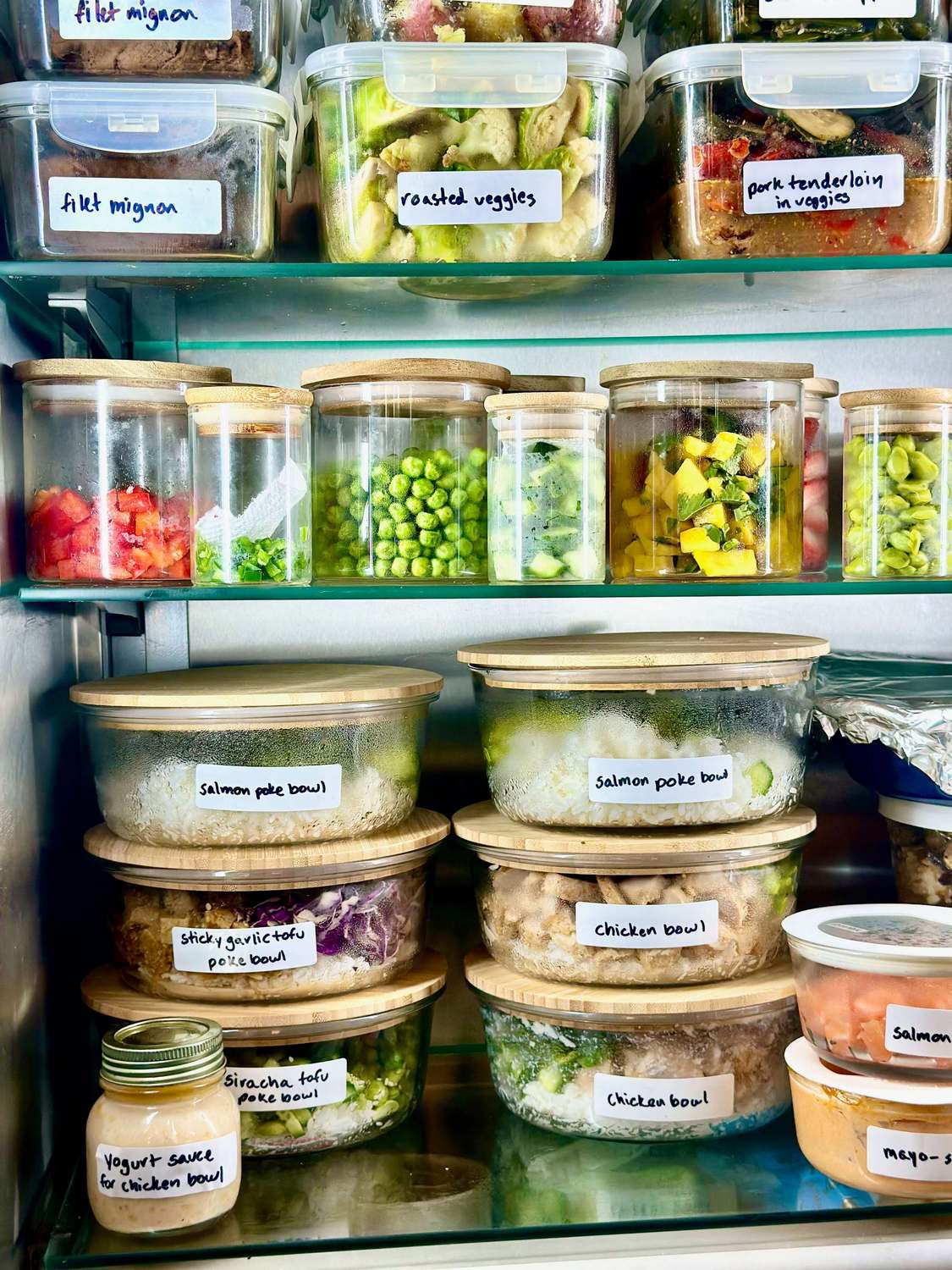 Refrigerator shelves holding labeled glass meal prep containers with bamboo lids, glass jars of prepped vegetables, and several plastic-lidded containers