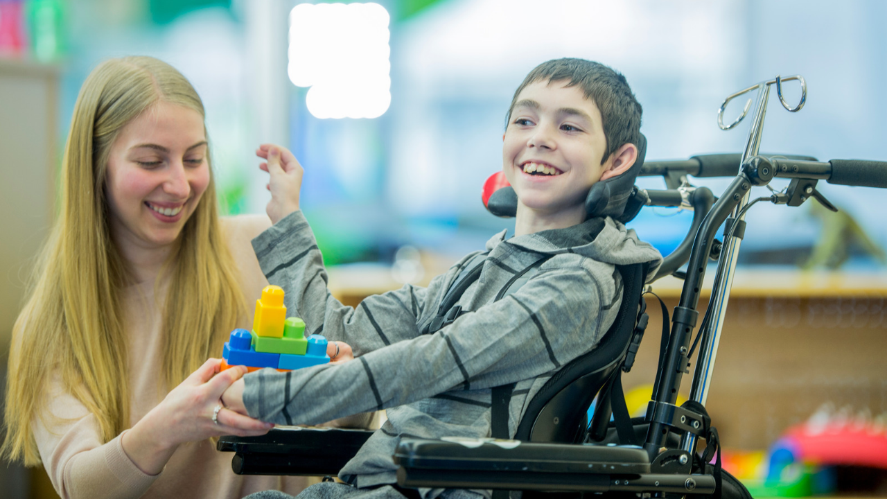 Therapist placing a block toy in the hands of a boy with cerebral palsy during physiotherapy to support grasp, hand control and movement skills.