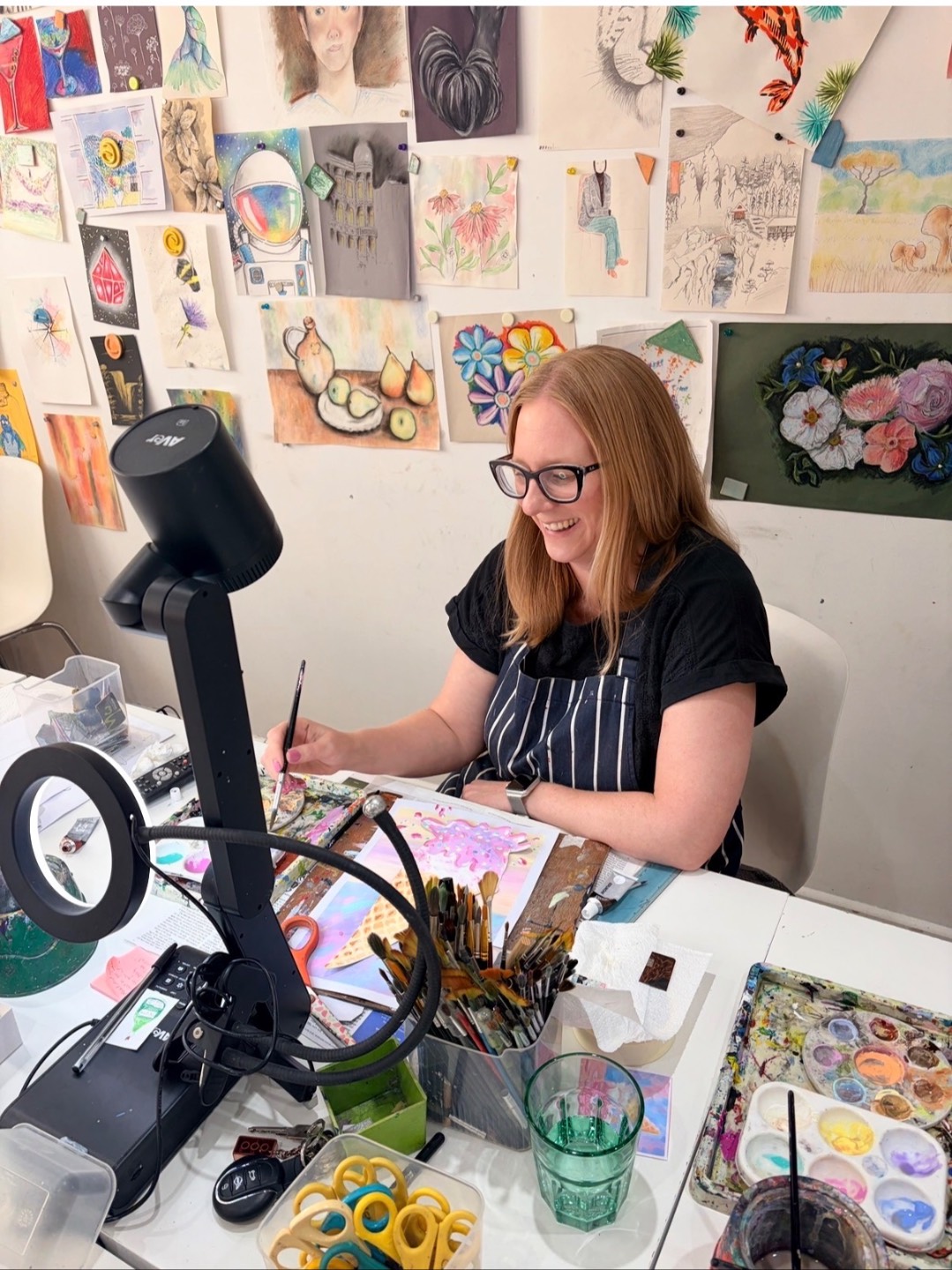 Helen Woodford smiling in the Create Art School studio, with art supplies laid out for a children’s painting class, ready to welcome young artists to a creative session.