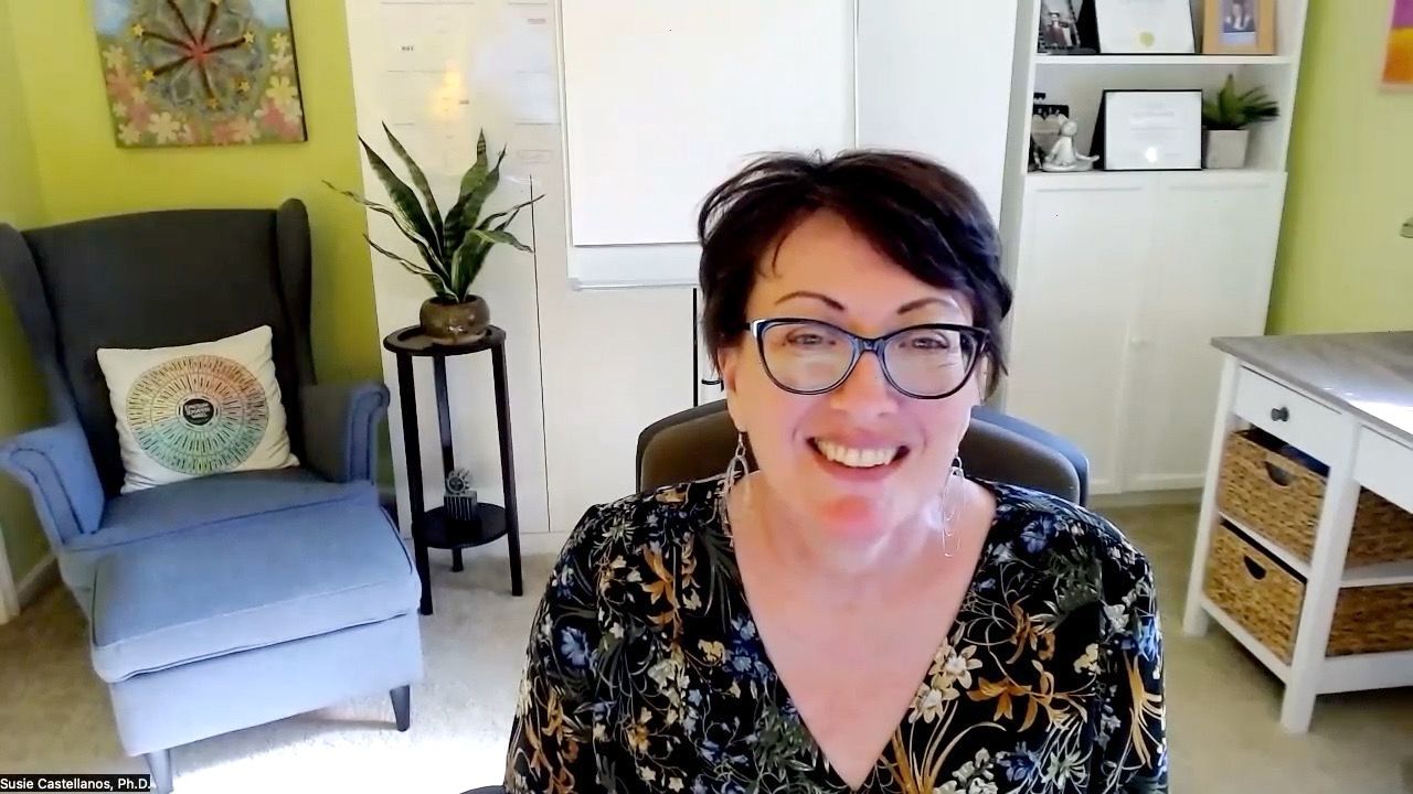 Photo of Susie smiling at the camera. The backdrop is her home office decorated in bright colors. She is wearing a black blouse with small blue and brown flower designs and is wearing glasses. 