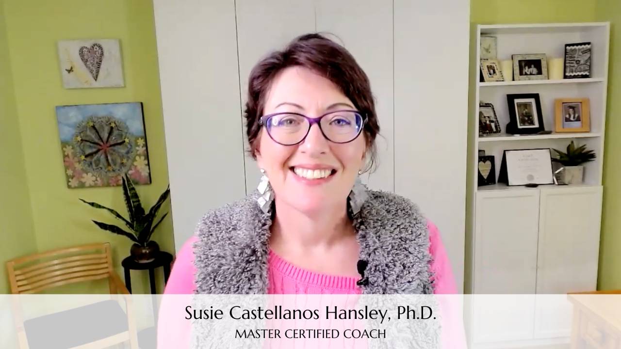 Photo of Susie smiling at the camera. The backdrop is her home office decorated in bright colors. She is wearing pink and gray and has glasses. Superimposed at the bottom of the image are the words "Susie Castellanos Hansley, Ph.D Master Certified Coach" on the bottom. 