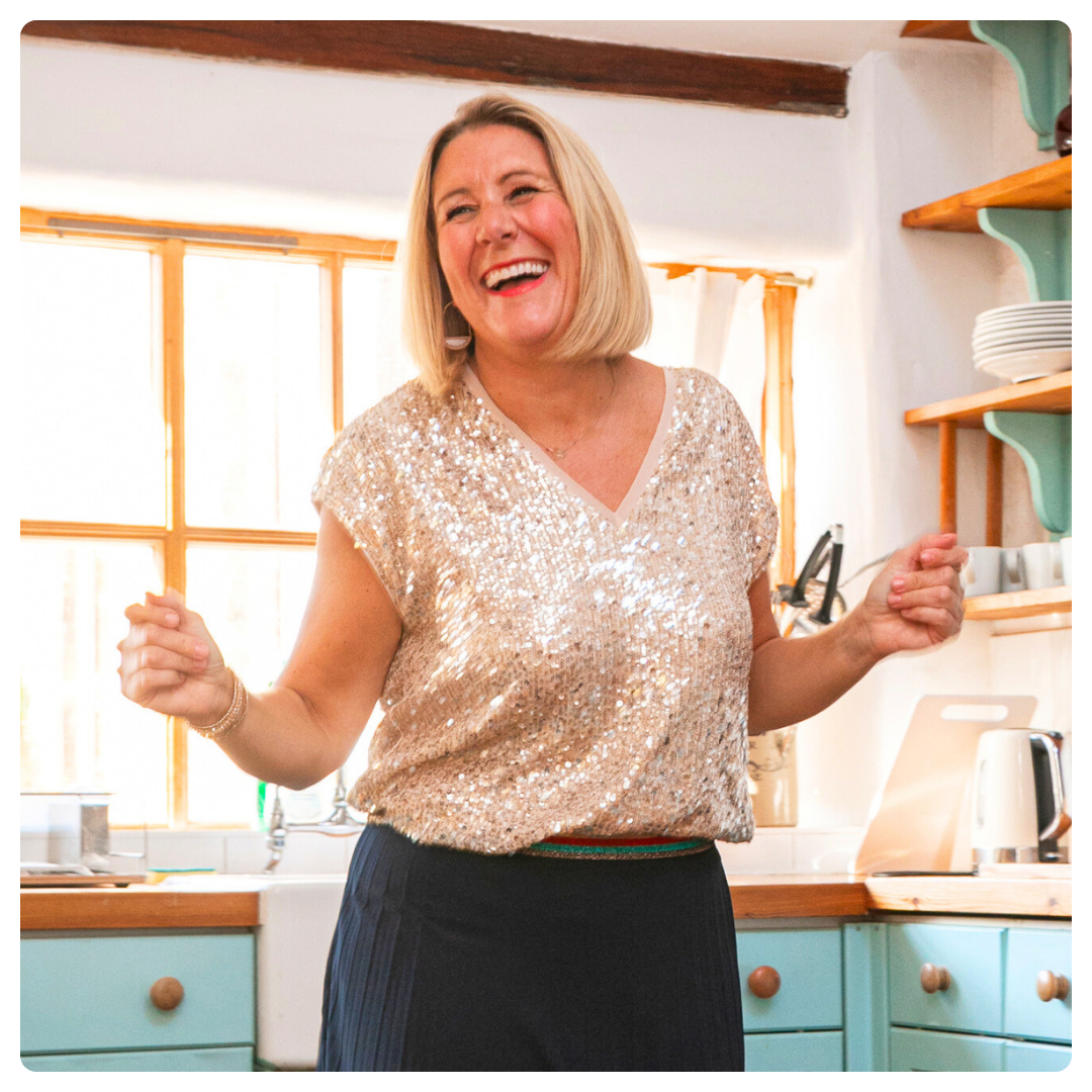 Karen Sutton sitting at a wooden table, smiling while holding a yellow mug. The table has a laptop, notebook, lemons, and a glass of water. The background features a teal couch, yellow flowers, and framed photos. The text below promotes a membership for widows to find community and healing.