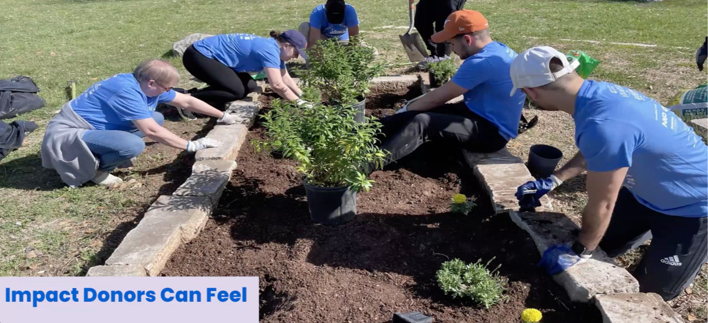 A group of volunteers in blue shirts work together outdoors to build a raised garden bed, placing stone borders and planting shrubs in soil, illustrating hands-on community impact and collective giving.