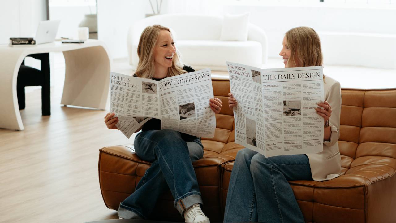 Krissy and Claire reading newspapers on a couch to symbolize them reading the AI Advantage Newsletter