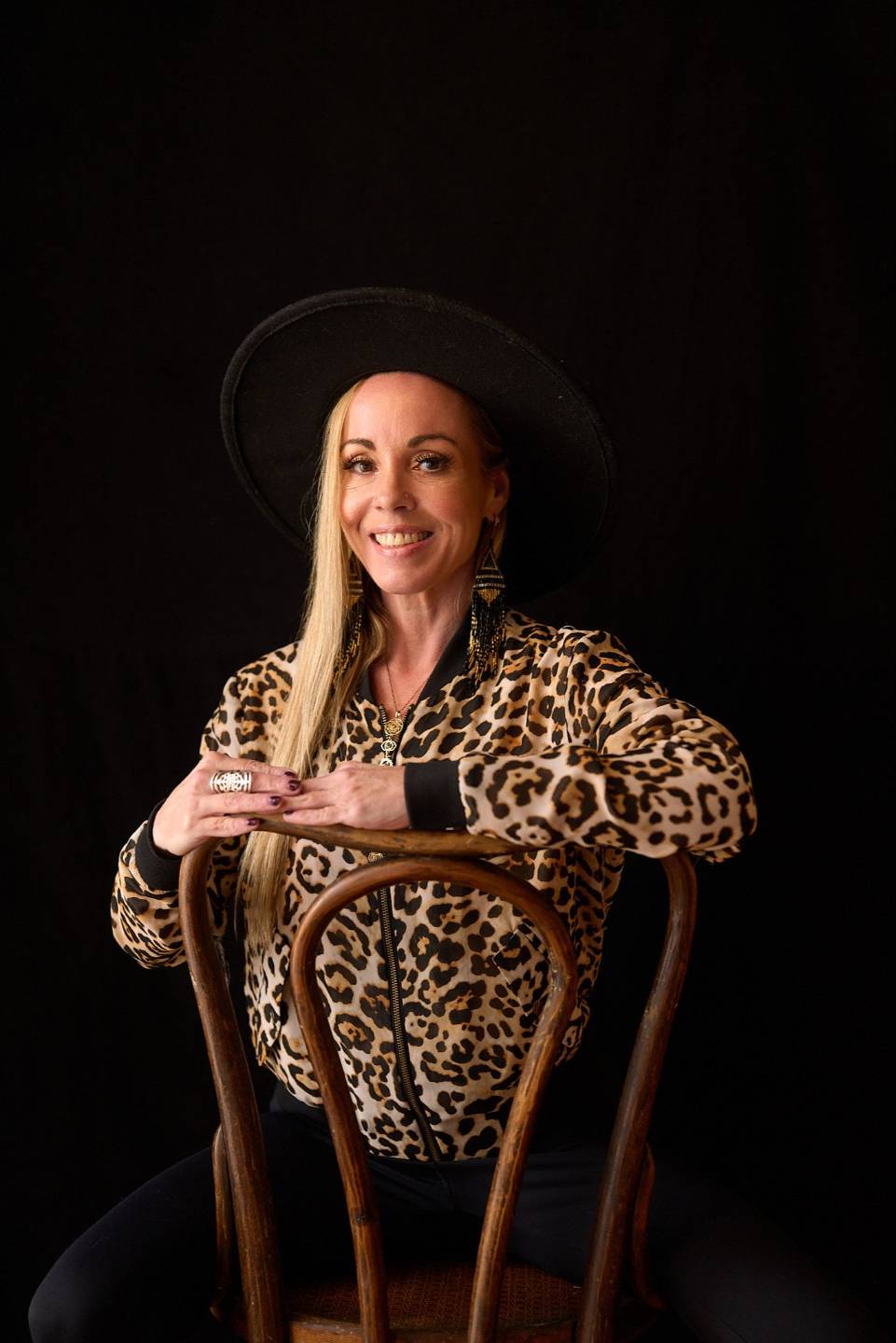 Dawn Light Amora, self love coach, in a leopard-print top and black hat, seated with arms folded over a chair