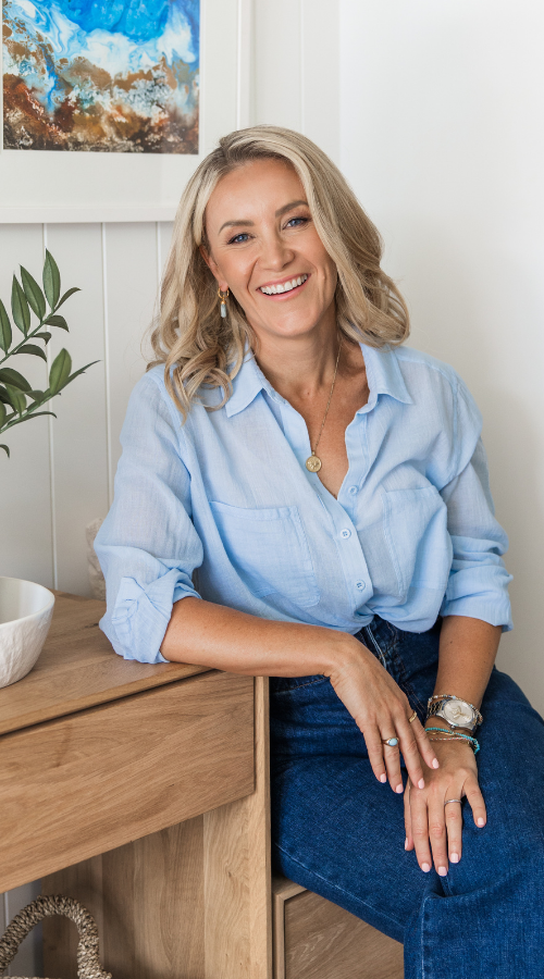 Denise Duffield-Thomas standing in front of timber desk with blue laptop in hand