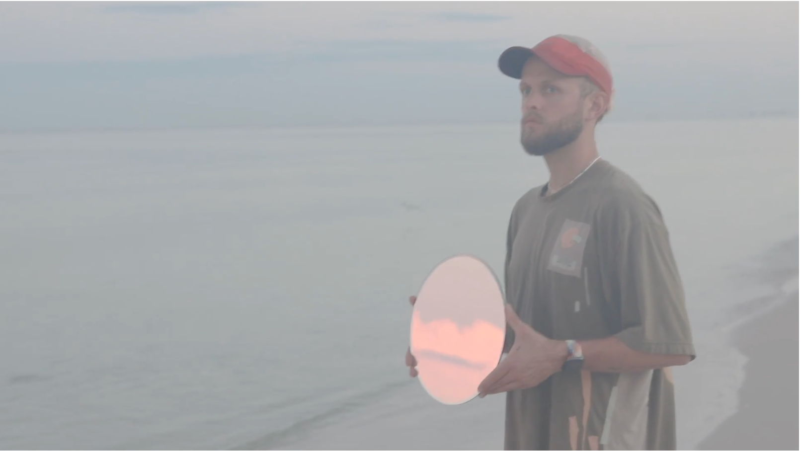 Rocco holding a mirror reflecting peach clouds beside the ocean