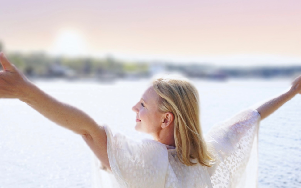 Woman standing with her arms raised, looking out over a lake, smiling in the sun.