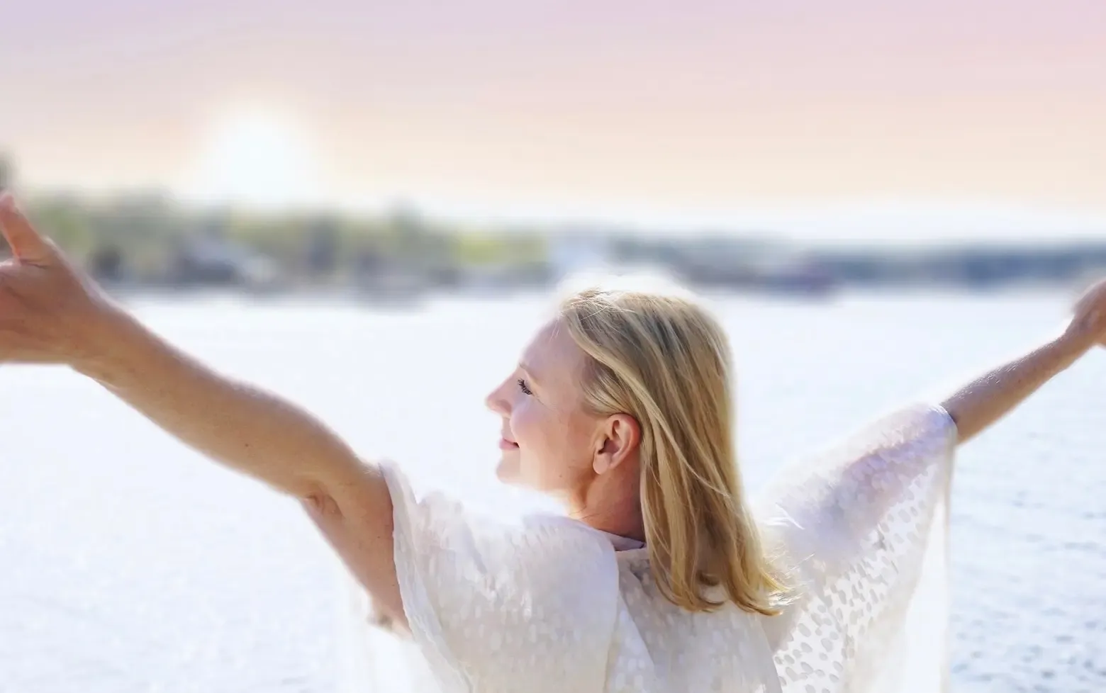 Woman standing with her arms raised, looking out over a lake, smiling in the sun.