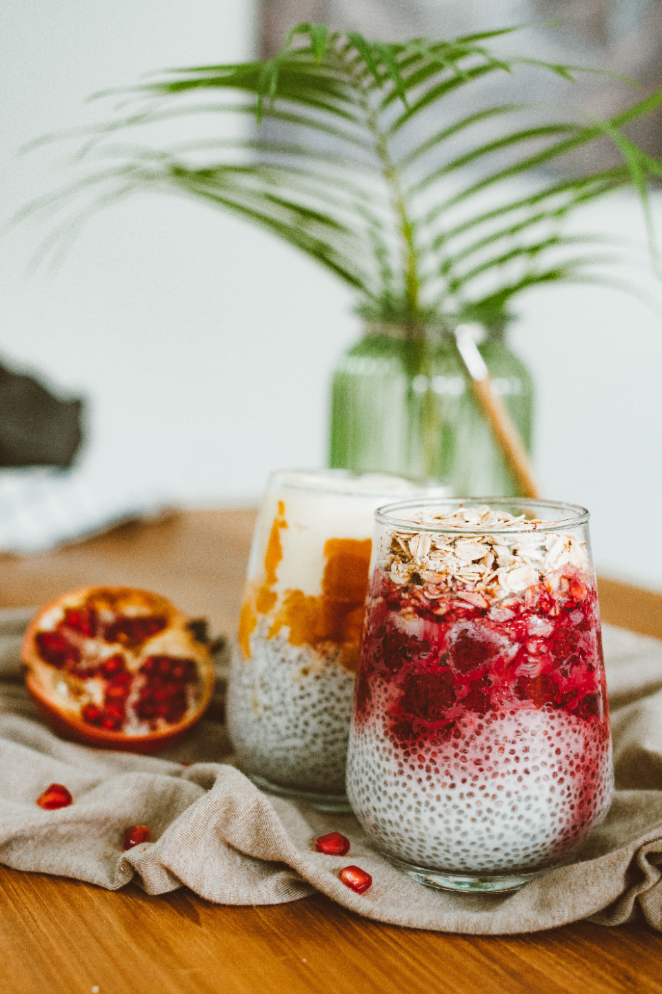 Two summer chia puddings with half a pomegranate beside. Sitting on a bench with a green fern in the background.