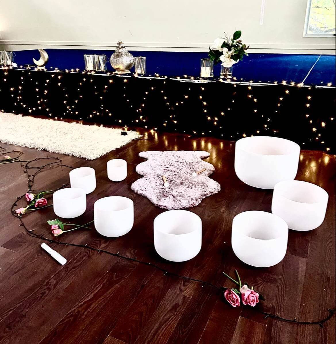 Crystal singing bowls arranged in a circle on a wooden floor with fairy lights, flowers, candles, and a decorated altar for a sound healing or meditation session.