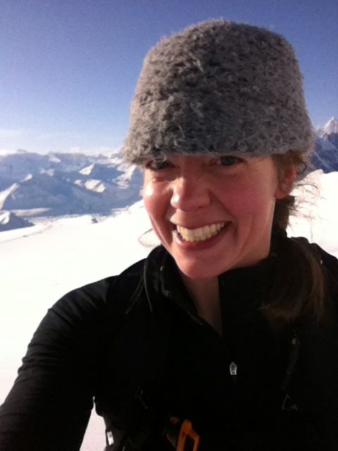 Shannon L. Post wearing a grey knit hat smiles brightly in a snowy mountain landscape, with clear blue skies and distant peaks behind her.