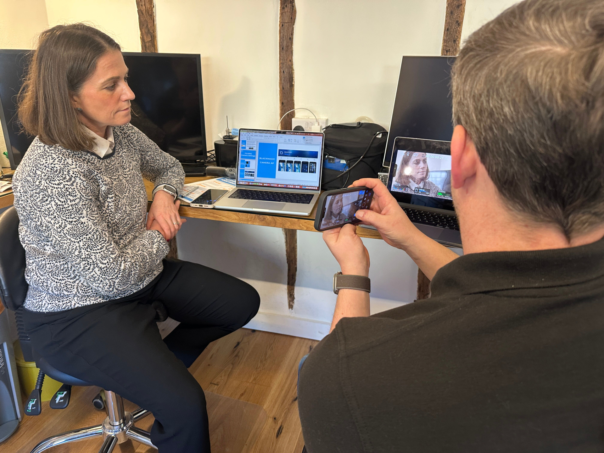 Tow people sitting with laptops and one is demonstraing how to use a mobile phone to film videos
