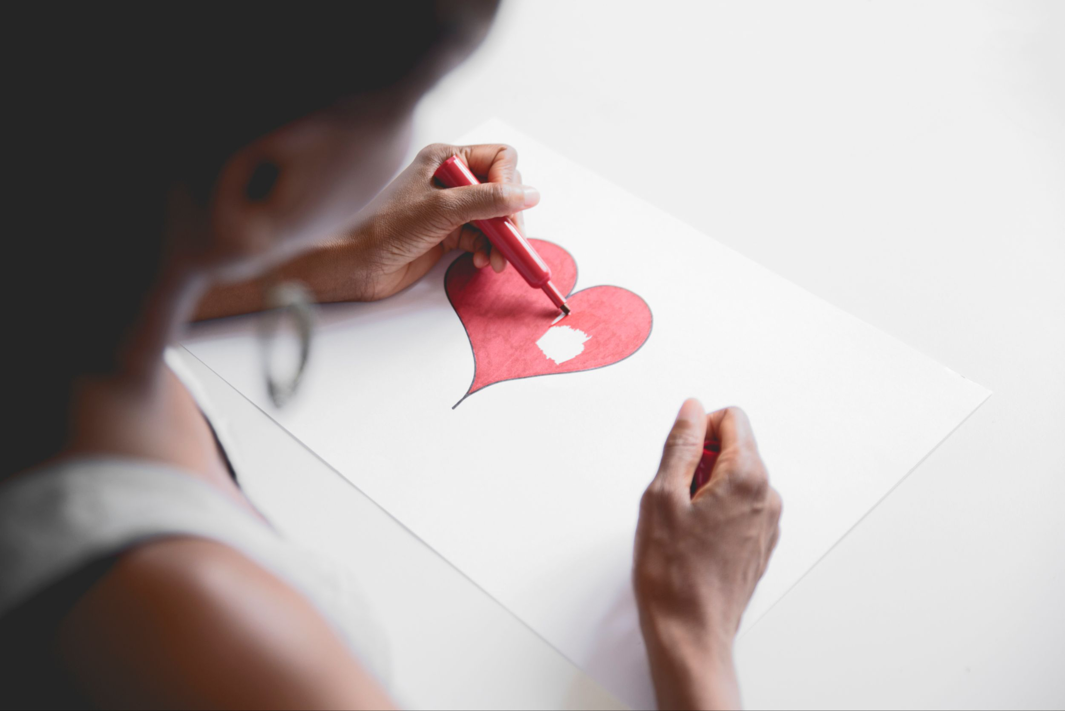 A black woman is wearing a grey tank top and has dangling hoop earrings. She is blurred out on the left side of the photo. In focus is her left hand holding a red marker as she colors a heart on a white piece of paper. 90% of the heart is colored. Her right hand rests on the piece of paper. The paper rests on top of a white desk.