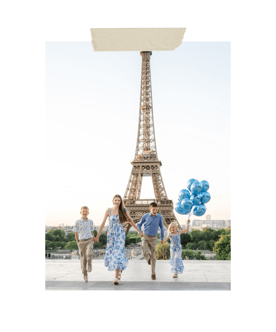 Family of four wearing coordinated blue and white outfits walking joyfully in front of the Eiffel Tower in Paris, with the youngest child holding blue balloons; bright morning sky and city view in the background.