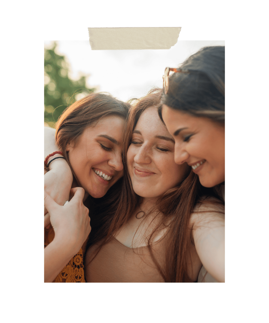 Three women embracing closely outdoors, smiling with eyes closed in a moment of warmth and connection, symbolizing friendship, community, and emotional support.