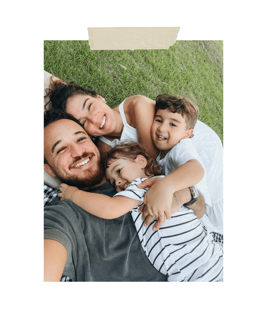 Smiling parents and two young children lying together on grass outdoors, hugging and enjoying quality family time in a candid lifestyle photo.